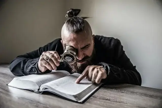 A photo of a man seated while reading his Bible with a magnifying glass signifying deep search.