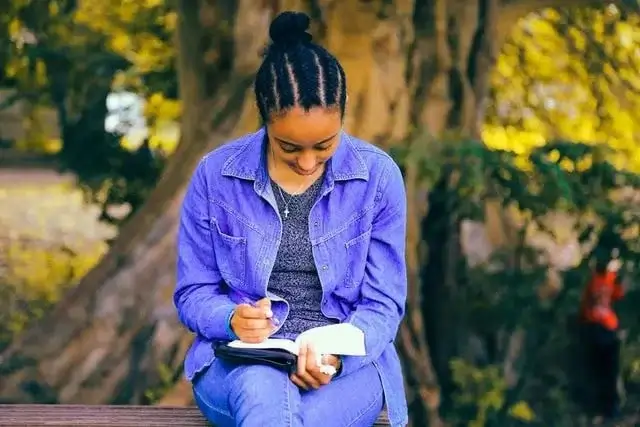 A photo of a young lady seated under a tree in an open ground studying the Bible