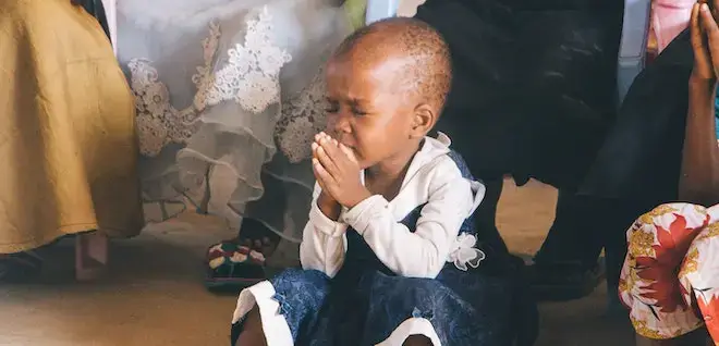 A small child with her hands clasped and eyes closed in a gesture of prayer in an African church