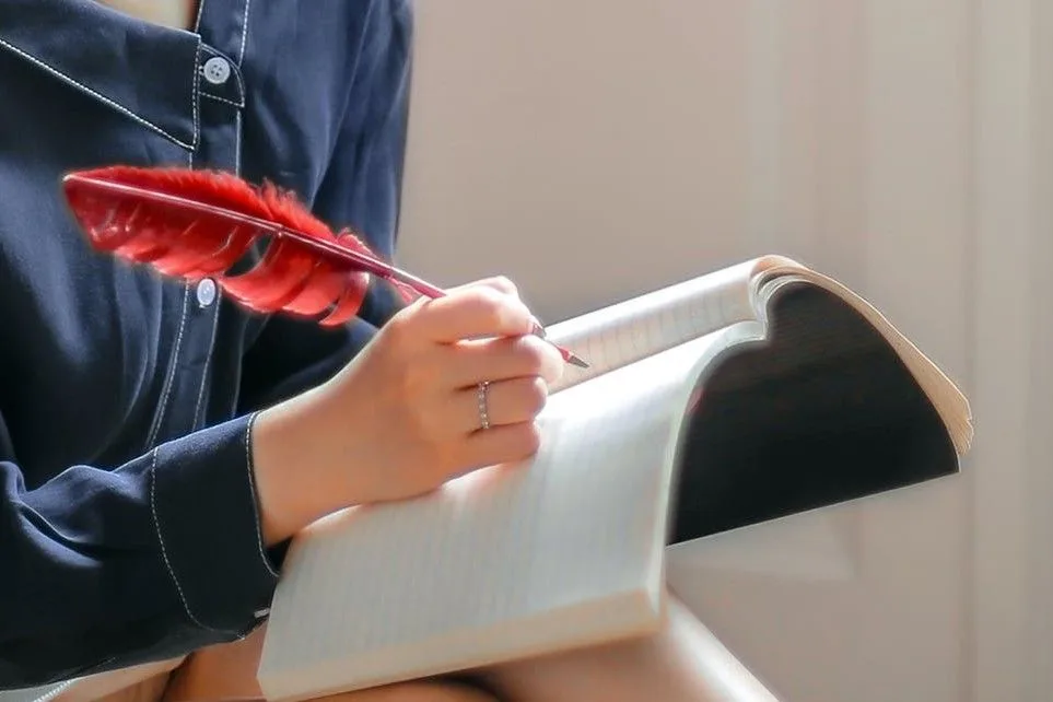 A photo of a ladies hand writing on a book with a red bird feather